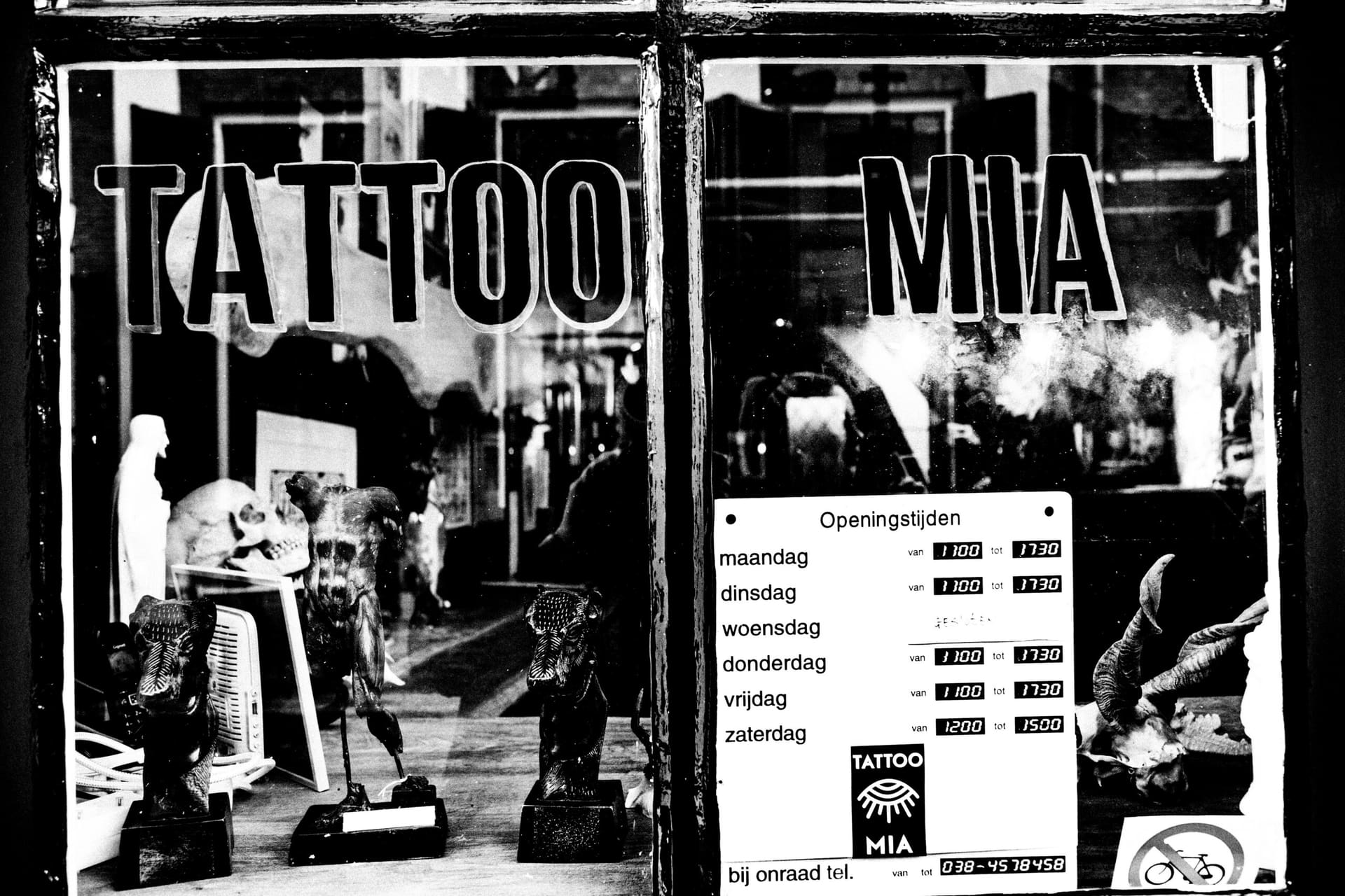 De etalage van de studio met de letters TATTOO MIA op het glas, beelden achter de ruit en het bordje met openingstijden.