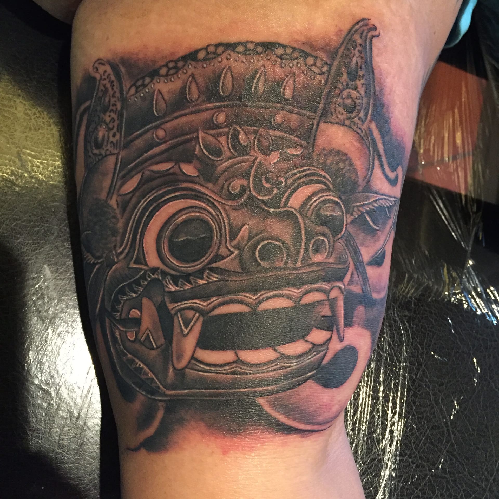 Tattoo van een Balinees Barong-masker in zwart en grijs op een bovenarm.